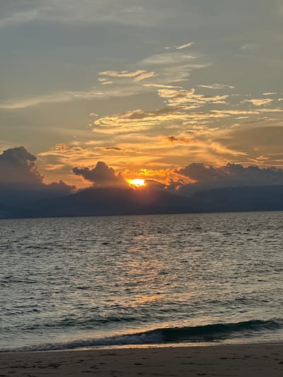 A sunset scene unfolds over the ocean with a shoreline in the foreground. The sun, partially obscured by dark, dramatic clouds and silhouetted mountains, casts a warm golden light across the sky and reflects on the water's surface. The ocean exhibits gentle waves lapping against a sandy beach, which shows faint footprints. The sky transitions from a pale blue at the top to shades of orange and yellow near the horizon, with scattered clouds catching the sunset's glow. This vista is characteristic of the coastal area of Saavedra, Philippines, known for its breathtaking sunsets over the water.