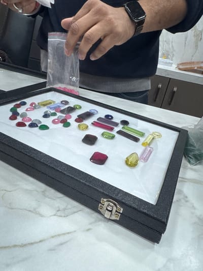 An elevated view captures a gemstone display inside a commercial establishment in Jaipur, India. The foreground features an open black display case containing approximately 60 polished, faceted gemstones of various shapes (oval, pear, cushion, emerald, baguette) and colors (red, blue, green, yellow, pink, purple, black) arranged on a white lining. A person, seen from the mid-torso, stands behind the case. The person's right hand holds an open clear plastic bag filled with small pink gems. The left hand displays a silver-colored ring and a black smartwatch. The individual wears a dark blue long-sleeved shirt over a grey underlayer and blue jeans. The scene occurs on a white marble-patterned countertop, with dark wooden cabinets and a light-colored wall visible in the background.
