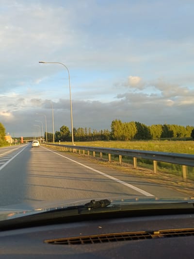 This media file shows a perspective from inside a vehicle traveling on a highway near Ādaži, Latvia. The scene is dominated by a highway with a metal guardrail on the right side. Several lampposts are lined up along the left side of the road, curving outwards towards the sky. The road appears to be asphalt, with white lane markings clearly visible. A white car is seen driving ahead on the highway.

The sky is a mix of blue and grey clouds, suggesting a dynamic weather condition, possibly transitioning from or towards rain. The lighting indicates it is either early morning or late afternoon, with the sun casting long shadows on the road. The right side of the image shows the interior of a vehicle, including a portion of the dashboard and a window, with some sort of vent visible on the far right.

The background features a line of trees and greenery, indicating a rural or semi-rural setting. The overall impression is one of travel and movement. There is no visible text in the image. The image is oriented vertically, suggesting it was captured with the camera held in a portrait orientation.