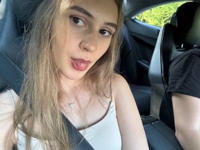 This is a close-up, sideways selfie of a young woman taken inside a car. The woman has long, blonde hair that partially obscures her face. She is wearing a light-colored top and a small, diamond pendant necklace. Her eyes are open, and her tongue is slightly sticking out. The background shows the interior of a car, including a seatbelt, and a glimpse of green trees and foliage through the window, suggesting an outdoor setting in daylight. The overall mood is casual and playful. There is no visible text in the image. The location context is Iffezheim, Germany.