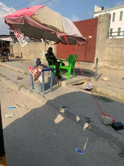 This image captures an outdoor, daytime scene on a street in Ikota, Nigeria, featuring a makeshift roadside service point. A large, weathered market umbrella with pink and beige panels provides shade over a blue metal table and two green plastic chairs. A single male individual, dressed in a dark t-shirt and trousers, is seated in one of the green plastic chairs, looking towards the left side of the frame with his right hand near his chin.
A white sign with red text, advertising "SERVICE," "MOBILE MONEY," "AIRTIME & DATA," and "MONEY TRANSFER," is affixed to the umbrella frame. A red rope extends from the umbrella, across the ground, and is tied to a large grey rock for stability. The ground consists of uneven dirt and concrete, with scattered debris including a blue wrapper and other small items. A concrete curb delineates the immediate area from a slightly darker, possibly paved, surface.
In the background, a concrete block wall runs horizontally, featuring a large red metal gate or door. Beyond this wall and gate, a multi-story white building with several rectangular windows is visible under a clear blue sky. Prominent shadows from the umbrella and other objects are cast on the ground, indicating direct sunlight.