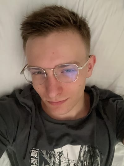 A person wearing glasses and a black t-shirt lies on a white bed. The person's face is turned towards the camera, with their head tilted. The t-shirt has a graphic design on it, including some text in what appears to be Japanese characters. The setting is indoors, likely a bedroom, with the person lying on a white sheet. The lighting suggests it could be daytime, but there are no clear indicators of the time of day or weather conditions. The person's expression is relaxed. The image is oriented sideways. The text on the shirt appears to be "破壊" which translates to "destruction" in Japanese.