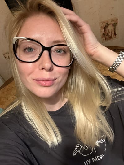A blonde-haired female individual is depicted in a close-up portrait, oriented forward, with light-colored eyes and a slight, closed-mouth smile. She wears black eyeglasses with white accents on the upper outer corners of the frames. Her left hand is raised to the right side of her head, with fingers partially obscured by her hair, appearing to adjust or rest in her hair. A silver-toned metal watch with a link bracelet is present on her left wrist. She is wearing a dark-colored, short-sleeved t-shirt with white lettering and a graphic design on the chest, including partial visibility of the word "selecty" and Cyrillic script. The background reveals an interior space with a wall featuring textured or tiled surfaces in warm, earthy tones. A small decorative tile or picture, depicting a scene with foliage and what appears to be bottles or figures, is visible on the upper right portion of the background wall. No specific architectural features or landmarks are discernible to forensically confirm the location as being in the city of Voskresensk, Russia.