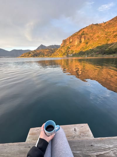 From a wooden dock in San Pablo La Laguna, Guatemala, a first-person view captures Lake Atitlán and surrounding mountains during golden hour. The lower body of an individual is visible, wearing light gray trousers. A left hand, adorned with a silver ring and light-colored nail polish, holds a light blue ceramic mug with dark patterns, filled with a dark liquid. The calm lake water reflects the warm, golden sunlight illuminating the rugged, vegetated slopes of the distant mountains and scattered shoreline structures. The sky exhibits partial cloud cover.