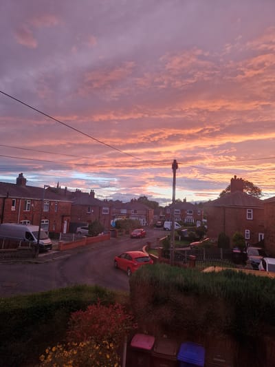 This is a high-angle, full shot of a street in Salford, United Kingdom, during a sunset. The sky is filled with dramatic clouds tinged with pink and orange. The street is lined with terraced brick houses. Several cars are visible on the street and in driveways, including a red hatchback, a silver van, and a dark-colored car. Power lines crisscross the sky, and a lamppost stands on the side of the road. In the foreground, a dense hedge partially obscures the view. There are no people visible in the scene.