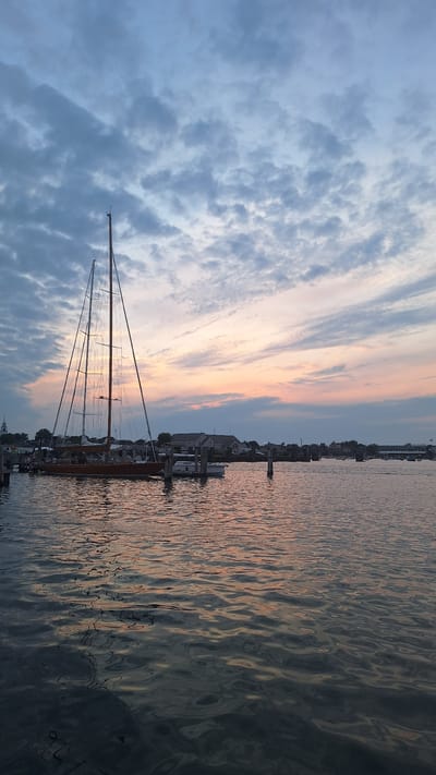 A panoramic view captures the sunset over Nantucket Harbor, USA, with a large sailboat prominently docked in the foreground. The sailboat, with its tall masts and wooden hull, rests on calm water that reflects the soft hues of the twilight sky. Several smaller boats are also moored nearby, alongside a pier. In the background, the Nantucket coastline is dotted with buildings and structures, including what appears to be a church steeple, bathed in the warm glow of the setting sun. The sky is a canvas of soft blues and pinks, streaked with clouds that are beginning to darken as night approaches. The overall atmosphere is serene and tranquil, conveying a peaceful end to the day.