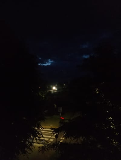 A nighttime, high-angle view shows a dark, cloudy sky with patches of lighter blue visible. In the foreground, silhouetted trees frame the scene. Below, a street scene unfolds, illuminated by artificial lights. A crosswalk with bright white stripes is visible on the asphalt. A traffic light with red signals is mounted on a pole. In the distance, streetlights cast a glow on the road and surrounding foliage. The overall impression is of a quiet, urban night in Kolno, Poland.
