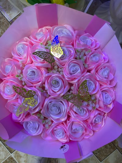 This is a close-up, top-down photograph of a bouquet of artificial pink and white roses. The roses appear to be made of satin ribbon and are arranged in a circular fashion. Several decorative gold and blue butterflies are interspersed among the flowers. Some of the roses have small clusters of white floral accents. The bouquet is wrapped in pink and white paper. A small "Thank You" sticker is visible on the lower left side of the wrapping. The background appears to be a tiled floor. There is no discernible indication of the location within the city of Armavir, Armenia, or any specific environmental context beyond what is directly visible.