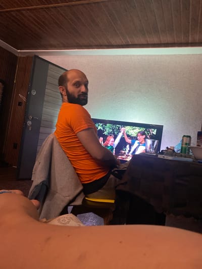 The image captures an indoor scene, likely a living room, from a slightly elevated, first-person perspective, with the photographer's bare arm and shoulder visible in the right foreground. A blue string or bracelet is on the arm, and part of a dollar bill is also visible.
The main subject is a man with a bald head and a beard, wearing an orange t-shirt and grey sweatpants. He is seated on a grey couch or armchair, his body angled towards a television screen, and he appears to be watching the content displayed.
The television, positioned centrally, shows what seems to be a show or movie. On the screen, two individuals are visible in an outdoor setting with lush greenery. One is a young woman with dark hair and glasses, wearing a white and blue top, depicted in a moment of apparent joy or excitement. Another person with red hair is partially visible at the bottom edge of the screen. A clear glass is resting on top of the television.
Behind the television, on a shelf or surface, various items are scattered, including several cans (one possibly a "Whiteclaw" product) and other unidentifiable objects. The room features a light-colored, textured wall on the right side and a dark wooden slatted wall or paneling on the left, separated by a thin vertical white strip. Below the TV, there's a dark piece of furniture with some circular details. The lighting suggests it might be evening or night, with the television screen illuminating parts of the scene. The overall impression is one of casual home entertainment.