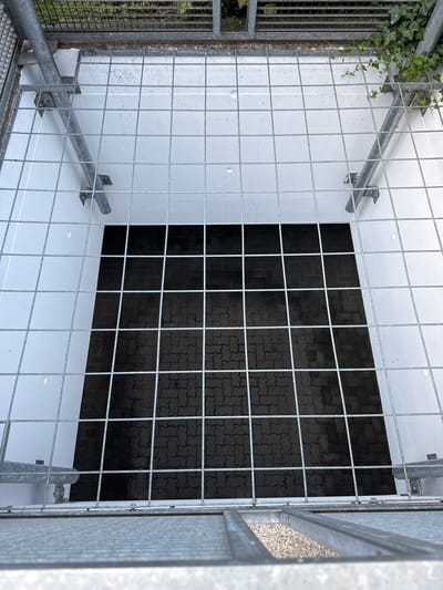 The image shows a top-down view into a caged area. The cage is constructed from metal grid fencing and has a white interior wall. Inside the cage, a section of dark grey interlocking pavers is visible. On the left side of the frame, some green ivy is growing on a metal grate. Metal supports and framing are visible throughout the cage, attaching the grid to the walls and providing structural integrity. The scene appears to be outdoors, possibly an industrial or utilitarian space, given the metal construction and the pavers. There are no people or animals visible. The lighting suggests it could be daytime, with no strong shadows indicating a specific time of day. The overall impression is of a secure or enclosed space, possibly a ventilation shaft, drainage area, or a secure enclosure for equipment. No text is visible in the image.