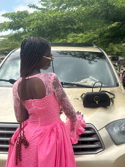 A young woman with braided hair, wearing a pink sequined dress and gold-rimmed glasses, stands with her back to the camera, leaning on the hood of a gold SUV parked outdoors. A black handbag rests on the hood. The scene is set against a backdrop of lush green trees and a cloudy sky. The location is identified as Itoga, Nigeria.
