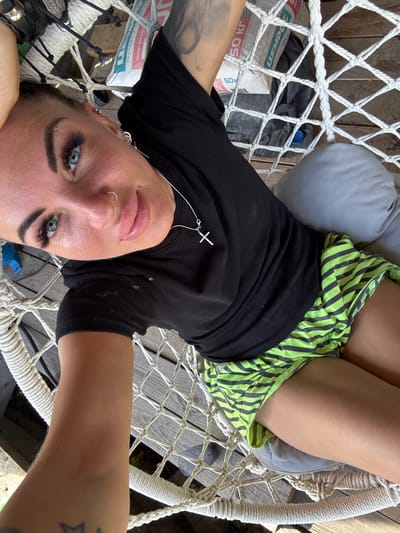 A woman is lying in a hammock, taking a selfie. The woman has long dark hair, blue eyes, and a nose ring. She is wearing a black t-shirt and green and black striped shorts. The hammock is made of white rope and is suspended from wooden beams. There is a gray pillow in the hammock. In the background, there are wooden boards and some bags. The lighting suggests it is daytime and sunny. The woman appears relaxed and happy. Visible text on a bag reads "50 КГ" and "УМЕНЬШЕНО".