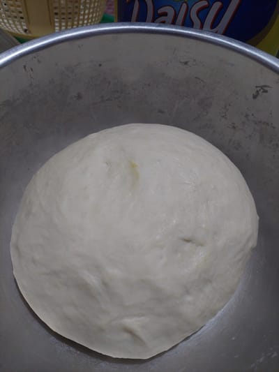 A close-up, overhead shot shows a mound of uncooked dough resting inside a stainless steel mixing bowl. The dough is smooth and pale yellow, with a slightly wrinkled texture. The bowl's rim is visible along the top and left edges of the frame, showing some wear and tear. In the background, the blurry edges of a yellow basket and a box with the "Daisy" logo are visible, suggesting a kitchen environment. The image appears to be taken in a residential setting in Jelutung, Indonesia, given the implied context of a domestic kitchen.