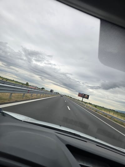 The image captures a view from inside a vehicle traveling on a highway in Malo Konare, Bulgaria. The sky is overcast with heavy, grey clouds. The highway is asphalt with clear white lane markings. On the left side of the road, a metal guardrail separates the highway from a dry, grassy field with sparse vegetation and a few distant trees. On the right side of the highway, there is a similar grassy field with more trees visible in the distance and a billboard advertising what appears to be a product with text that reads "Nuii". Several vehicles, including cars and trucks, are visible on the highway ahead, suggesting moderate traffic. The foreground shows the dashboard and windshield interior of the vehicle.