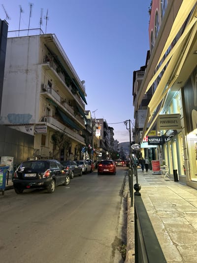 This image depicts an urban street scene in Koridallos, Greece, captured during early evening or dusk. The clear sky overhead transitions from a soft blue to a deeper twilight hue. The view looks down a street lined with multi-story buildings on both sides, characteristic of an urban environment.
On the left side of the street, buildings feature shops on the ground floor, identifiable by their awnings and illuminated interiors, with residential units occupying the upper levels. Balconies with greenery and numerous television antennas on the rooftops are visible. The right side of the street also has buildings, some with ground-level businesses and apartments above.
The paved street accommodates both parked and moving vehicles. A line of cars, primarily compact sedans and hatchbacks, are parked along the right side of the road, with one black car displaying the license plate "ZKB 1108." Further down, a red car is seen in motion, indicating ongoing traffic. A sidewalk runs alongside the right-hand buildings, separated from the road by a black metal railing. A few pedestrians are discernible on the sidewalks. Streetlights are illuminated, casting a warm light on the scene.
Visible text on shop signs includes "PORTOBELLO'S," "V STYLE," "CAFE MEAL," and "ESSY." Greek text, such as "ΓΙΑΛΟΜΑΤΩΝ" (likely a business name) and possibly "ΠΩΛΟΥΝΤΑΙ" (meaning "for sale") on a banner, can also be observed on buildings and signs. The scene conveys the atmosphere of a lively neighborhood as evening settles.