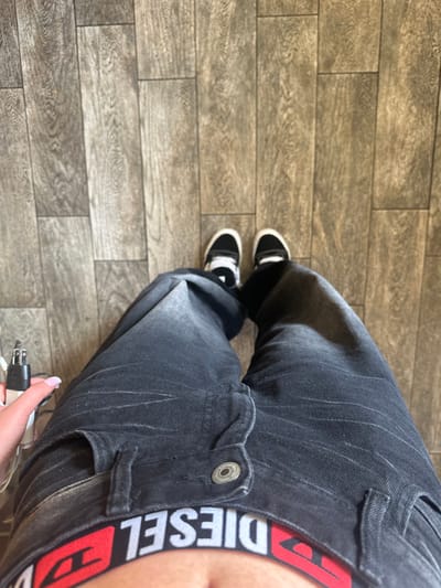 The image captures a first-person, downward-facing view of an individual's lower torso, legs, and feet within an indoor setting in Glendale, United States. The specific location within Glendale is not visually identifiable from the image. The individual is wearing dark gray, faded denim jeans with a visible metal button closure on the fly. A red and white elastic waistband, displaying the reversed text "DIESEL," is exposed above the jeans. On the feet are black low-top sneakers with white soles and laces, worn with white socks. The floor beneath is constructed from rectangular planks exhibiting a light brown wood-grain pattern, consistent with laminate or tile flooring. The individual's left hand, featuring light pink manicured fingernails, is holding a white electronic device, identified as a power adapter or charger, which has a two-prong (Type A US standard) plug at its top and a black cable extending from it.
