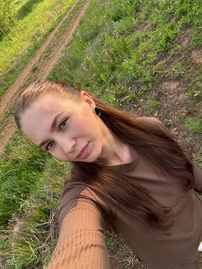 A young woman is captured in a close-up selfie or portrait, looking directly at the camera with a calm expression and a slight smile. Her head is tilted, giving the image a Dutch angle. She has long, straight, dark brown hair that cascades over her shoulder and is also visible spread around her on the ground. Her eyes are a striking green, and she has well-defined eyebrows and eyelashes. On her visible ear, she wears a black circular earring with a gold rim.

She is dressed in a brown, long-sleeved top or dress with a distinct ribbed texture. The setting is outdoors, likely a rural or semi-rural area in Nizhnyaya Gar', Russia. The background consists predominantly of vibrant green grass and patches of dry earth. A dirt path or track is visible curving through the grassy landscape, suggesting an open field or trail. The lighting appears to be natural daylight, indicating either morning or late afternoon, creating a soft glow on her skin and hair. There is no visible text in the image.