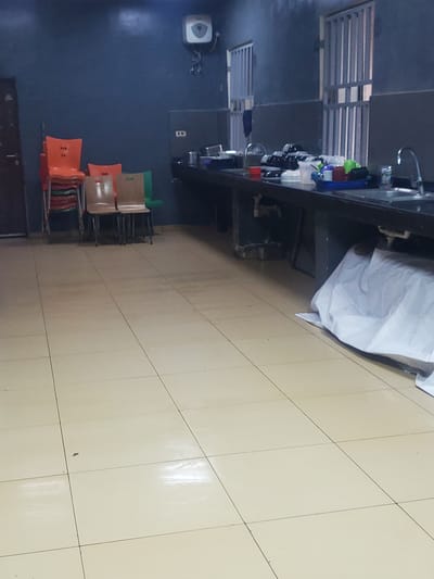 The image depicts a dimly lit room, possibly a communal kitchen or dining area, located in Gwagwada, Nigeria. The floor is tiled with light-colored squares, showing some wear and tear. To the left, a stack of orange chairs is visible, with a few wooden chairs placed beside them. A dark-colored door is partially visible on the far left.
The main focus of the scene is a long, dark countertop, likely a kitchen counter, running across the middle and right side of the image. It is cluttered with various kitchen items. On the counter, there are stacks of plates, cups, and other dishes, some appearing to be clean and neatly arranged, while others are in a blue basin, suggesting they are in use or waiting to be washed. A faucet and sink are visible on the right side of the counter. Behind the counter, a dark wall with barred windows suggests the building might be secure.
A large white sheet or cloth is draped over a section of the counter or furniture underneath it on the right side, partially obscuring what is beneath. In the upper left corner of the image, a white unit, possibly a water heater or air conditioning unit, is mounted on the dark blue wall. The overall impression is one of a functional, albeit somewhat unkempt, common area.