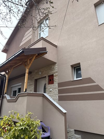A detailed forensic description of a residential exterior in Kumanovo, North Macedonia, captured during daylight hours.

The scene depicts the facade of a two-story building featuring a textured, light beige stucco finish. The upper section includes dark brown eaves. On the upper right, a rectangular window is equipped with a partially raised white exterior roller shutter. Below this, a smaller, recessed rectangular window with white trim is positioned above two horizontal, dark brown decorative stripes on the wall, which terminate in angled shapes.

The main entrance is covered by a wooden canopy, supported by two light-colored wooden posts with dark metal bases, and topped with a dark brown metal roof and gutter system. The wall surrounding the entrance is faced with light-colored stone veneer. A white entrance door with an oval glass insert is visible. To the immediate right of the door, a small, rectangular red box (likely a mailbox or utility access) is affixed to the stone veneer.

An outdoor staircase, constructed with a beige stucco base and capped with white marble-like accents, ascends to the main entrance. Below the main staircase, on the right, a separate set of grey, granular steps descends towards a lower ground level. A window with a fully lowered white exterior roller shutter is located on the lower left portion of the building.

In the lower foreground, a green and yellow variegated shrub is prominently featured, partially obscuring a purple plastic outdoor chair positioned against the building wall. Bare tree branches with visible green buds extend into the upper left and central portions of the image, indicating early spring. A dark utility cable traverses the upper left section of the frame. No individuals are present in the scene.