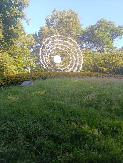 A large, metallic, spiral-shaped sculpture is situated in a grassy area. The sculpture, which appears to be an abstract form, is comprised of interconnected metallic elements and has a central spherical component. It is surrounded by lush green grass and a line of yellow-flowering bushes. In the background, a dense arrangement of trees forms a backdrop, with the clear blue sky visible above. A small, grey, angular object, possibly a concrete or stone structure, is partially visible in the grass to the left of the sculpture. The scene suggests an outdoor public space, potentially a park or garden. The image was taken in Jasseron, France.