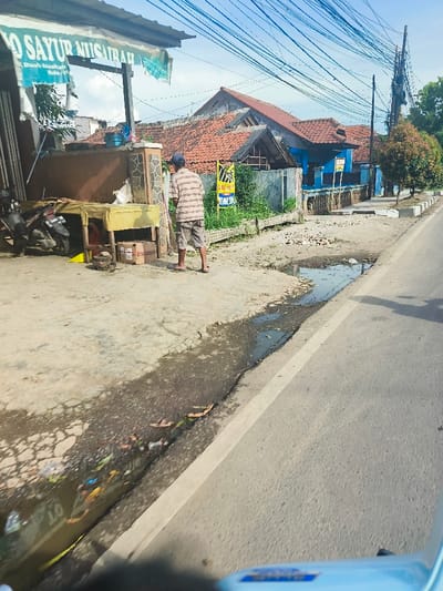 The scene is taken from a street-level perspective, looking toward the right, capturing a man standing by the side of a road in Purwakarta, Indonesia, at the coordinates -6.552, 107.437. The man is wearing a striped shirt and pants, and is facing towards the right. He is standing in front of a small shop with a sign that reads "SAYUR NUSANTARA". There are various small objects and plants in the area around the man. The road is paved and has a white painted lane divider. It is mostly clear, with a small puddle of water to the left of the lane divider. The surface to the left of the road is uneven and covered in dirt and mud. It is overgrown with small plants and debris. There is a small wooden table with some unknown objects sitting on it. There are several power lines running across the sky, and a few trees and houses are visible in the background. The buildings are mostly single-story and made of brick. The sky is blue with a few clouds. The scene is taken in daylight.