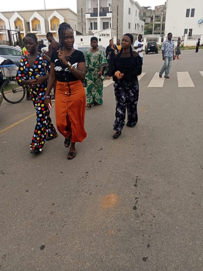 In Guzape, Nigeria, a group of individuals is captured in an outdoor scene, possibly a street or a residential area, with modern buildings in the background. The foreground is dominated by a paved road, with visible white pedestrian crossing lines, indicating a crosswalk.
Four women are the central focus of the image. The woman on the far left is wearing a colorful, polka-dot patterned dress, featuring a vibrant mix of blue, red, yellow, and white dots against a black background. She has dark hair styled in braids and appears to be looking downwards. To her right, a woman in a black top with a lace-up detail and an orange, high-waisted skirt with prominent buttons is walking. Her hair is also styled in braids, and she is looking forward. A third woman, dressed in a green and white patterned gown, is walking in the middle ground, looking to her left. She has dark hair and appears to be wearing glasses. The woman on the far right is wearing a black top and black patterned pants, with a black headscarf. She is also looking forward.
Other individuals are present in the background, including a man in a patterned shirt and jeans walking across the crosswalk, and a man in a white t-shirt and dark pants. A black car is parked in the middle ground, and to the left, a bicycle with a basket is visible. The surrounding environment features contemporary architecture, with buildings showcasing a mix of white and grey facades, and some structures appearing to be under construction. The overall atmosphere suggests a daytime scene in an urban or semi-urban setting.