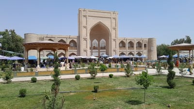 The image depicts a bustling outdoor market scene in front of a large, ancient-looking beige brick building with arched doorways and windows. The building appears to be a historical monument or religious site. In the foreground, a manicured lawn with scattered evergreen shrubs and trees is visible. A pathway runs through the lawn, bordered by a decorative fence.
The market itself is set up with numerous stalls covered by colorful tents, primarily blue and white, displaying various goods. People are seen browsing, shopping, and interacting within the market area. Some individuals are wearing traditional Uzbek clothing. To the left, a wooden gazebo with an ornate roof and columns offers shade.
The overall environment is sunny with a clear blue sky. Trees line the perimeter of the market and the historical building. The architecture of the building suggests a historical significance, possibly a madrasah or mosque, characteristic of the region of Denov, Uzbekistan. The presence of market stalls indicates a vibrant local economy and cultural activity.