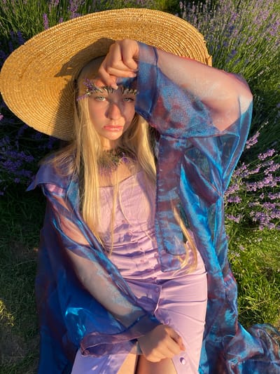 A young woman with long blonde hair lies on her back in a field of lavender. She is wearing a light purple dress and a shimmering blue and purple sheer scarf draped over her. A straw hat is positioned near her head. Lavender sprigs are tucked around her face and in her hair. The woman is looking up at the camera with her left hand near her face. The scene is illuminated by bright sunlight, suggesting it is daytime. The lush green grass and blooming lavender indicate a warm, pleasant weather condition and a rural or natural setting. The overall mood appears serene and picturesque. The location is Verkhneye Uchdere, Russia. There is no visible text in the image.
