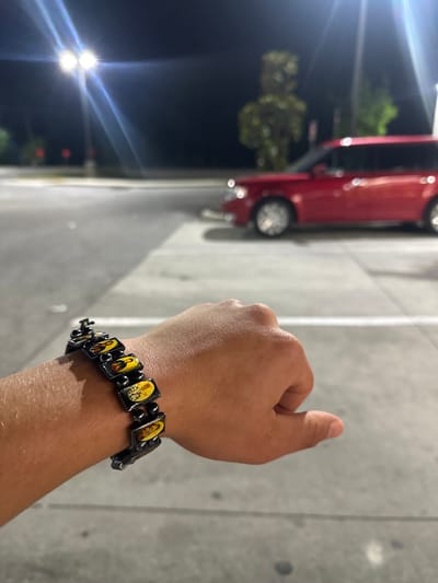 A close-up, outdoor shot shows a person's right hand and wrist with a black bracelet featuring small, rectangular images of religious figures against a yellow background. The person's thumb is extended upwards. In the blurred background, a red vehicle is visible, with a dark sky, streetlights, and greenery further in the background. The location cannot be determined as Homosassa Springs.