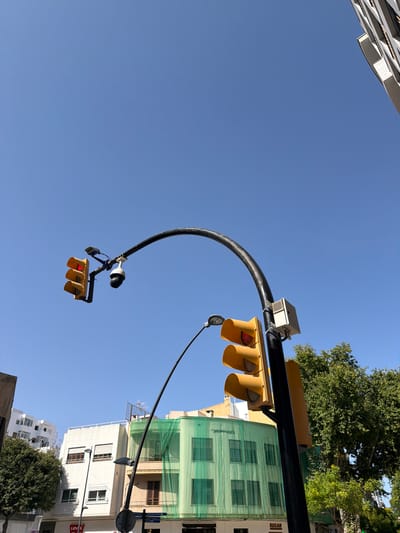 This is a photograph taken from a street level perspective, looking up at a bright, clear blue sky in Ibiza, Spain. The image is dominated by a traffic light with a red light illuminated, mounted on a curved black pole that extends across the frame. Attached to the pole, below the traffic light, is a black and white surveillance camera. Further to the right, a second traffic light, also yellow, is visible, with its lower lights illuminated. This second traffic light is mounted on a black pole that extends vertically. Behind the traffic lights and poles, several buildings are visible. To the left, a white building with multiple windows and balconies stands out. In the center, a building is covered in green netting, suggesting construction or renovation. To the right, a large, leafy green tree occupies a significant portion of the frame. The overall impression is an urban street scene on a sunny day.