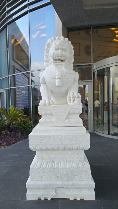 A white marble statue of a Foo dog, a mythical lion-like creature, stands imposingly on a multi-tiered pedestal in front of a modern building with large glass windows and a revolving glass door. The statue faces the viewer directly, its mouth open and mane detailed. The pedestal is intricately carved with decorative patterns. To the left of the statue, lush green palm plants and purple foliage are visible. The ground is paved with dark grey tiles. In the background, through the glass, a person can be seen walking through the revolving door into the building, and reflections of the interior are visible in the glass facade. The overall scene suggests an entrance to a contemporary establishment in a city, possibly Agios Epiktitos, Northern Cyprus / Cyprus, given the architectural style and landscaping.