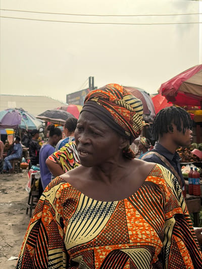 The image captures a bustling open-air market scene in Jakande, Nigeria, under an overcast sky. In the foreground, an older woman with dark skin is prominently featured, looking towards the left with her mouth slightly open, appearing to be speaking or reacting. She wears a vibrant, patterned garment in shades of orange, yellow, and black, and a coordinating headwrap. Her hair is covered by a dark fabric under the patterned wrap.
Behind her, the market extends into the middle and background. To her immediate right, a younger man with dreadlocks is visible in profile, looking towards the right. He wears a dark shirt. Adjacent to him is a stall featuring produce, possibly green watermelons, and several plastic bottles, likely containing beverages.
Further in the background, numerous people are present, moving or standing. Several market stalls are visible, characterized by various colored umbrellas, including red, light blue, and darker hues, providing shade. Corrugated metal roofs are discernable behind these stalls. The ground appears to be unpaved, dusty earth. Overhead, power lines span across the frame. A large billboard or sign is visible in the distant background, partially obscured. The overall environment suggests a lively, active market setting.