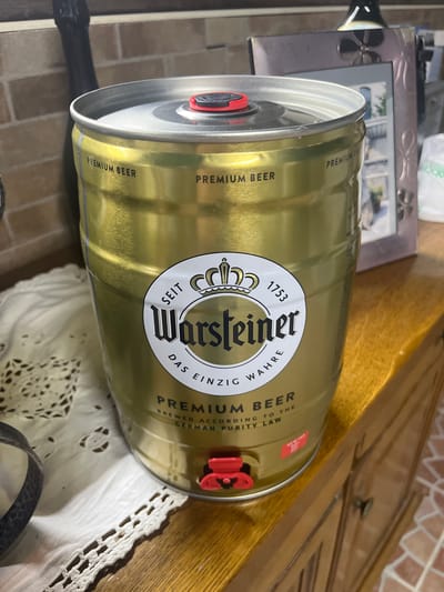 The image shows a cylindrical keg of Warsteiner Premium Beer. The keg is gold-colored with black lettering, and has a red dispensing tap. The text on the keg reads "Warsteiner" in large letters, along with "Premium Beer", "Das einzig wahre" (The only true), and "Brewed according to the German purity law". The number 1753 is also visible. The keg is positioned on a surface that appears to be a combination of a wooden table, a white lace tablecloth, and a brick-like surface. A silver picture frame is also partially visible in the background. There is no indication of the location being in Ceraso, Italy.