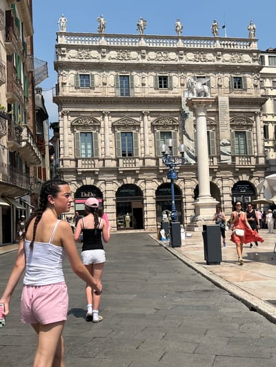 The image shows a street scene in Verona, Italy, dominated by a tall, ornate building with classical architecture. The building features multiple stories with windows and statues along the top. A tall white column stands in front of the building. There are two women in the foreground, one wearing a white top and pink striped shorts, the other in a black top and white shorts. They are walking on a stone-paved street. In the background, other pedestrians can be seen. Storefronts line the ground floor of the building, including a luxury eyewear store and a clothing boutique called "DoDo". A sign on the column lists several famous artists. The sky is clear and blue.