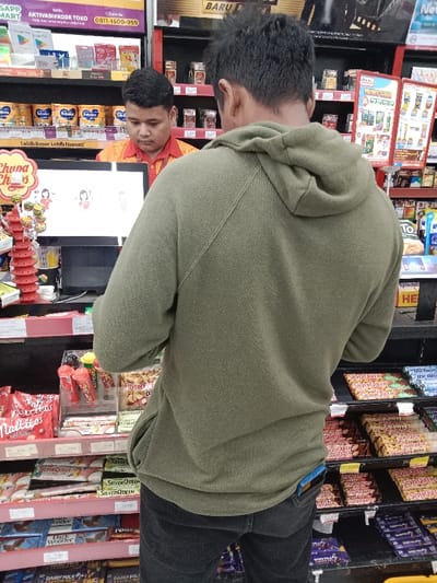 The image shows a young man wearing a green hoodie and black jeans standing in front of a cashier in a convenience store in Kaliwungu, Indonesia, located at coordinates -6.953, 110.239. The man is facing the cashier and appears to be looking at the cashier. There is a computer monitor in front of the cashier and the man is slightly out of focus. There are various snack foods on the shelves behind the man and the cashier. The shelves are red and black. There are various candy and snack items on the counter. There are some items displayed on a red stand next to the computer monitor. The store has a black and red color scheme.