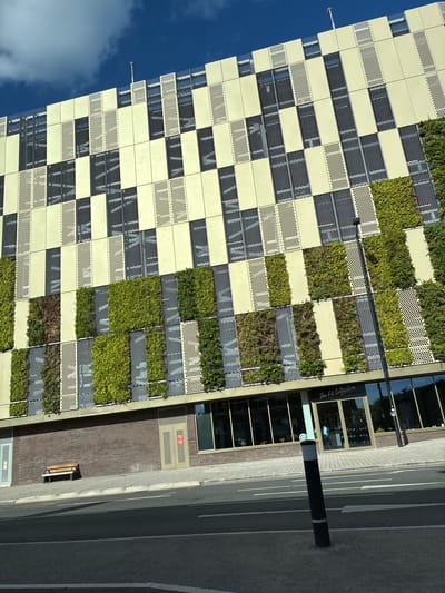 The image shows an exterior view of a modern building with a distinctive facade in Chester, United Kingdom. The building features a grid-like design with alternating sections of pale yellow panels, dark grey mesh screens, and vertical green walls. To the right of the building, a storefront with the name "The Fit Collective" is visible. A black bollard stands on the sidewalk next to a paved brick area. The street, marked with white lines, is partially visible. A bench and a fire exit door are also visible next to the building. A section of blue sky and white clouds is visible in the upper left corner of the frame.