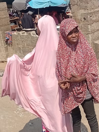 A woman wearing a red and white patterned hijab and black pants is seen smiling in the image. She is lying down, making a gesture with her hands. Beside her is another woman covered in a light pink hijab and gown. In the background, there are clothes hanging on a line and a brick wall. The picture is taken outdoors with bright sunlight.