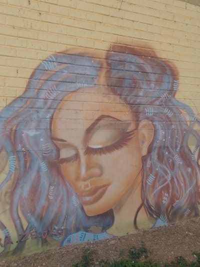 The image shows a mural painted on a yellow brick wall. The mural is a portrait of a woman with her eyes closed, depicted in profile. She has long, flowing hair with blue and pink highlights, adorned with decorative patterns that resemble stylized sound waves or DNA strands. The woman's face is rendered in soft, muted tones of peach and brown. Her eyelashes are long and emphasized. The background wall has a textured, brick pattern. The mural appears to be located outdoors, near ground level, with a dirt and gravel embankment in the foreground. Some greenery is visible on the embankment. Faint graffiti, possibly a tag or artist's signature, is visible at the bottom of the mural. The overall aesthetic is artistic and somewhat ethereal. The graffiti itself, a stylized portrait, is the primary subject. There is no specific indication of human activity beyond the creation of the mural. The environment suggests an urban setting, likely an exterior wall exposed to the elements.