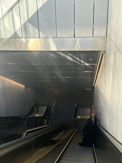 An indoor scene shows a person riding an escalator in a modern building. The walls are lined with rectangular panels in light and dark metallic hues. The escalator has a black handrail and steps with a visible yellow striped pattern. The overall aesthetic is clean and minimalist, with light playing off the metallic surfaces.