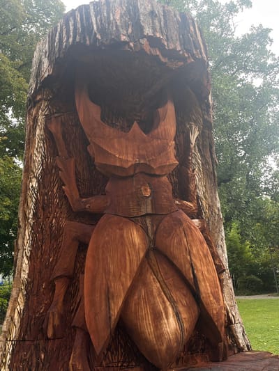 The image presents a large, vertical wood carving of a stag beetle, located outdoors in a public green space within La Tour-de-Peilz, Switzerland. The sculpture is carved directly into a substantial tree trunk or stump, with the rough texture of the original tree bark retained on the upper and outer edges of the work. The central focus is the brown-toned wooden beetle, depicted with its distinctive large, curved mandibles at the top, a segmented body, and multiple legs carved in relief against the trunk. The surface of the carving displays visible wood grain and tooling marks. The environment includes a grassy area in the foreground and background, with other mature trees visible behind the sculpture under what appears to be an overcast sky. No individuals or active interactions are depicted in the scene.