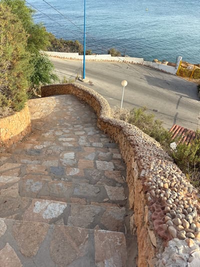 The image shows a stone staircase descending towards a paved road that curves alongside the Mediterranean Sea in Urbanización Cabo Roig, Spain. The staircase is bordered by a low, curved stone wall topped with small, rounded pebbles and a larger, rougher stone wall. Lush green vegetation grows to the left of the staircase, and sparser, dry brush and some red-tiled roofing are visible to the right. A blue pole stands near the road, which is illuminated by a white lamppost. The sea is a clear blue, with a small yellow buoy visible in the distance. The overall scene suggests a scenic coastal pathway.