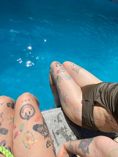 The image shows two individuals sitting by the edge of a swimming pool. Both individuals have multiple colorful tattoos on their legs. The person on the left has tattoos of a smiley face, a geometric shape, flowers, a zebra, and other abstract designs. The person on the right has tattoos of a spider, flowers, a tiger's face, and abstract lines. The pool water is a bright blue and reflects the sunlight. The individuals are sitting on the edge of the pool, which appears to be made of stone or concrete. The environment is sunny and outdoors. The image does not contain any specific information that would suggest the location is in Mtskheta, Georgia.