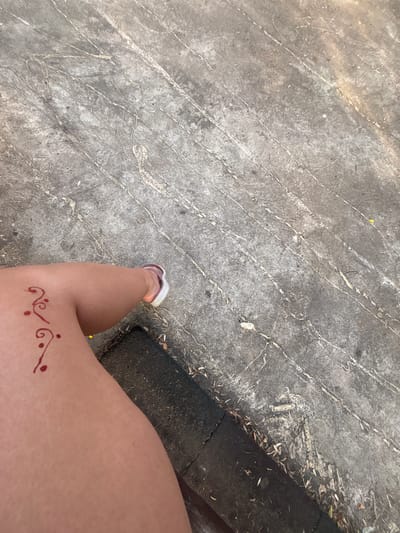 The image shows a person's left leg with a dark red henna design on the thigh. The design is abstract, with swirls and dots. The person is wearing a white sandal with a dark strap. The leg is positioned on a textured gray surface that appears to be concrete or asphalt. The background shows more of the same textured surface, with some debris and dried leaves scattered around. The lighting suggests it is daytime, with sunlight casting shadows. The location is identified as Garmen, Bulgaria.