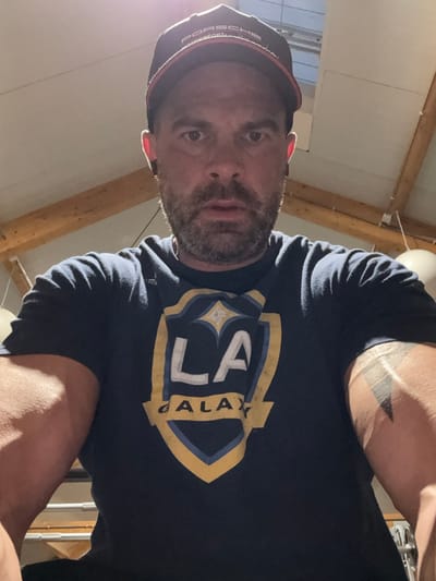 An adult male subject, appearing to be in his 30s to 40s, is depicted in a close-up, head-on portrait, likely a self-taken photograph. He possesses short dark hair, a stubble beard, and dark eyes, staring directly into the camera. He wears a black baseball cap with a red brim, displaying "PORSCHE MOTORSPORT" text on the front. Black in-ear devices are visible in both ears. He is dressed in a dark blue short-sleeved t-shirt featuring a prominent "LA GALAXY" shield logo, which includes "LA" in white, "GALAXY" in gold, and a gold and blue shield design with a star motif. A dark, geometric tattoo, composed of stacked triangular shapes, is visible on his right upper arm. The background reveals an indoor setting with a light-colored, possibly panelled, ceiling intersected by exposed wooden beams. A bright light source, consistent with a skylight, is present in the upper center, casting a visible lens flare. A spherical, light-colored object is partially visible on the right side of the background. The scene is located in Jemelle, Belgium.