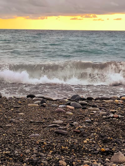 The image depicts a rocky beach with the sea and a cloudy sky. The foreground is filled with an abundance of dark, smooth pebbles of various sizes, interspersed with smaller stones and some lighter-colored, smooth stones. A wave is breaking in the middle ground, with white foam crashing onto the shore. The sea is a choppy, dark blue-gray color. The horizon line separates the sea from the sky, which is dominated by a band of bright yellow and orange, suggesting either sunrise or sunset, with dark, heavy clouds above. The scene is devoid of any people, structures, or vegetation. The location is identified as Zemo Kvariati, Georgia, suggesting a coastal setting in that region.