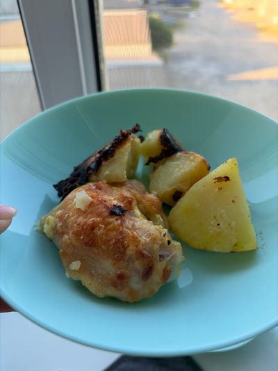 A close-up, overhead shot shows a light blue plate of food, likely a meal, placed near a window. The plate contains a piece of golden-brown, crispy chicken and several chunks of roasted potatoes, some with slightly charred edges. The background is blurred, showing a window with a view of buildings and trees. The overall impression is of a simple, home-cooked meal. The image's metadata indicates it was taken in Kaspiysk, Russia.