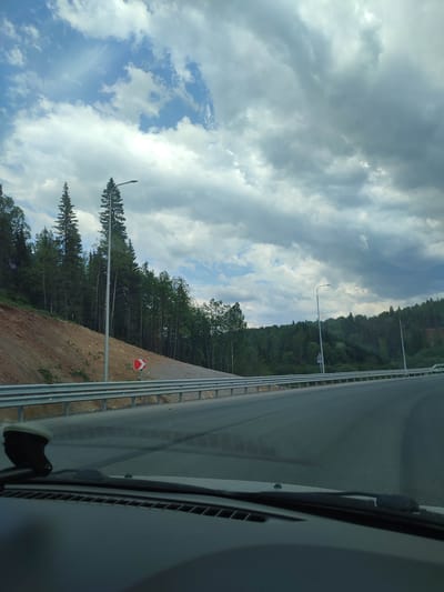 The image, likely taken from inside a vehicle, shows a view of a road in a wooded area near Gremyachinsk, Russia. The sky is cloudy with a mix of blue and gray patches. The road curves to the right. A metal guardrail runs along the side of the road, protecting a bare dirt slope with some trees. A road sign indicating a curve is visible. Streetlights are present along the roadside. The dashboard of the vehicle is in the foreground, with a windshield wiper and a mounting device. The general environment is rural with a mix of coniferous and deciduous trees. No people are visible.