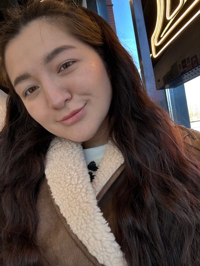 A woman with long brown hair is wearing a brown jacket with a white fur collar. She is indoors. The image has a sign in the background. The city and country is not determinable from the image.