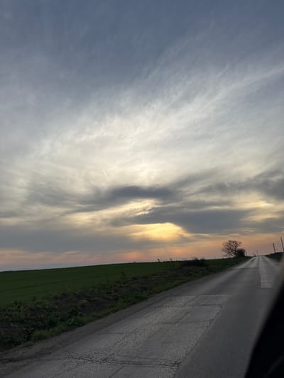 The image shows an asphalt road running along a green field in Sofronievo, Bulgaria. The sky is filled with wispy, grey and white clouds, with some areas showing a golden glow. A faint contrail is visible in the sky. A tree with bare branches stands near the road. Power lines run alongside the road. The image is taken from a slightly elevated position.