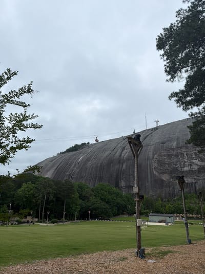 The image captures Stone Mountain in Stone Mountain, Georgia, United States. The large granite dome dominates the frame, its textured gray surface rising sharply. A cable car system ascends the mountain, with one car visible on the cables. Metal towers support the cables along the mountainside. Green grass covers the area at the base of the mountain, bordered by a gravel pathway. Trees line the edge of the green space. The sky is overcast, casting a diffuse light over the scene.